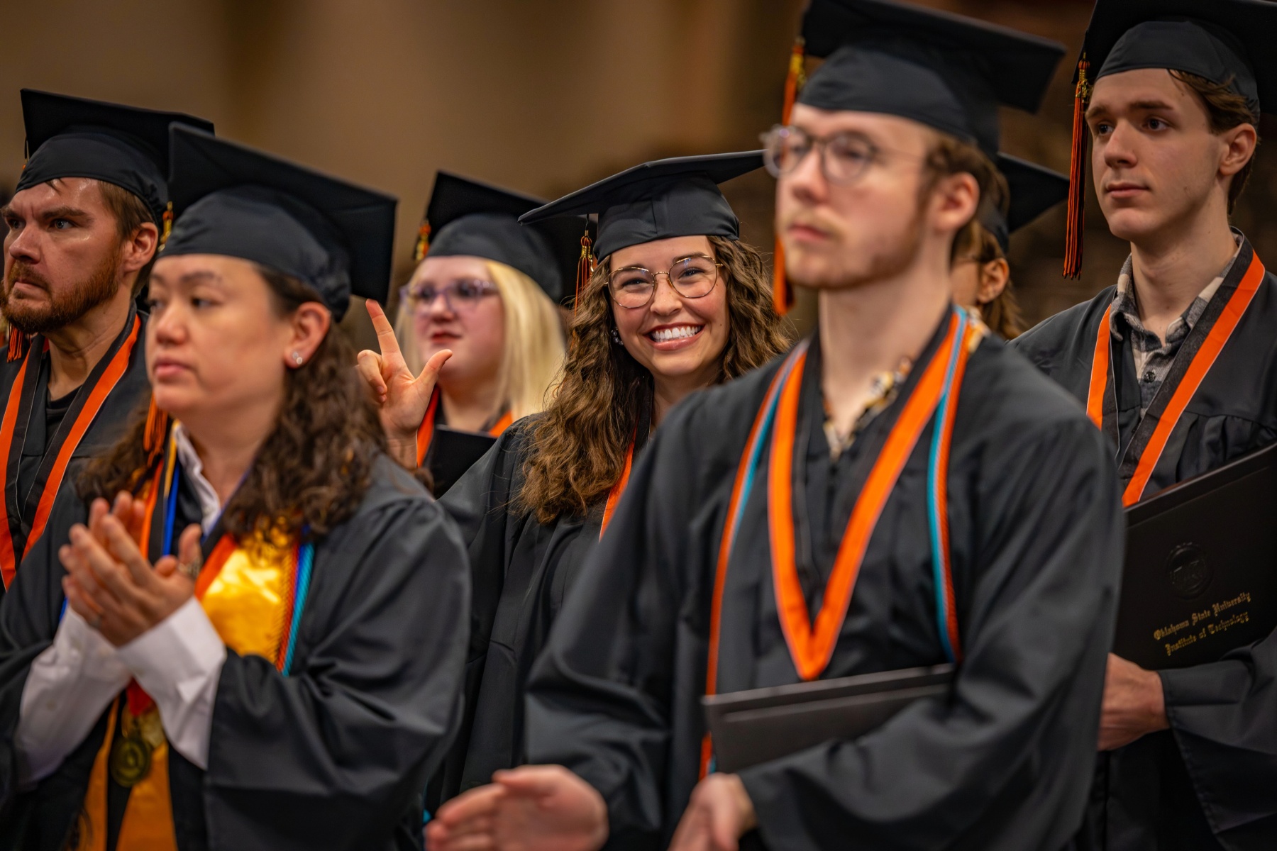 Graduates wearing black caps and gowns sit together during an OSUIT commencement ceremony, applauding and smiling as the ceremony continues.