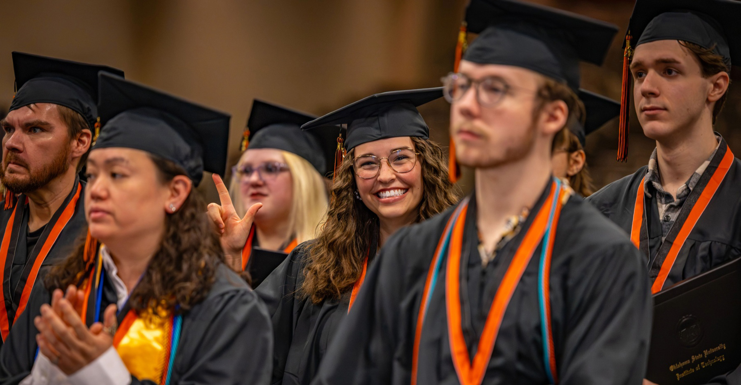 Graduates wearing black caps and gowns sit together during an OSUIT commencement ceremony, applauding and smiling as the ceremony continues.