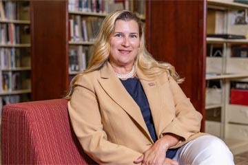 A headshot of Dr. Cynthia Hatch, an OSUIT social sciences professor, smiling in front of a background of library bookshelves filled with books. She has blonde hair and is wearing a tan blazer over a dark top, a pearl necklace, and a small flag lapel