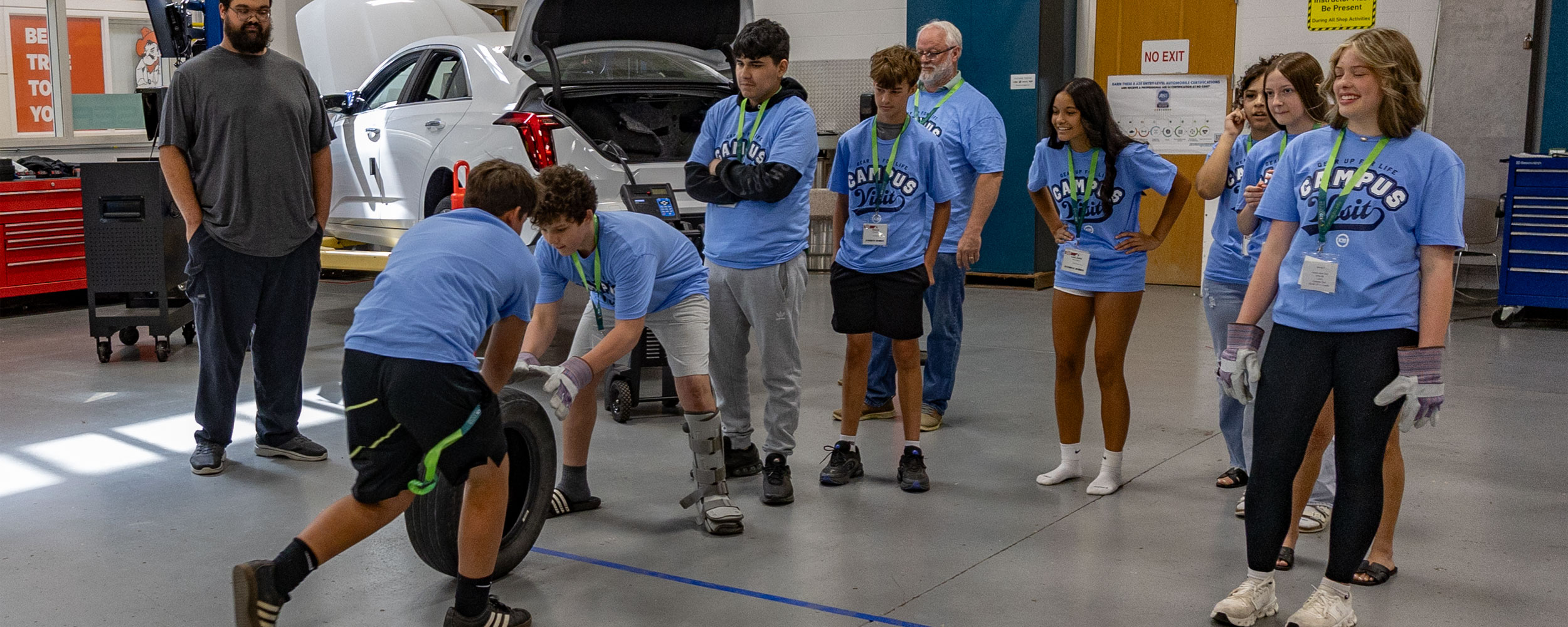 A group of students wearing matching “Campus Visit” T‑shirts watch and participate in a hands-on activity in an automotive lab, where two students roll a tire while a car sits on a lift in the background.