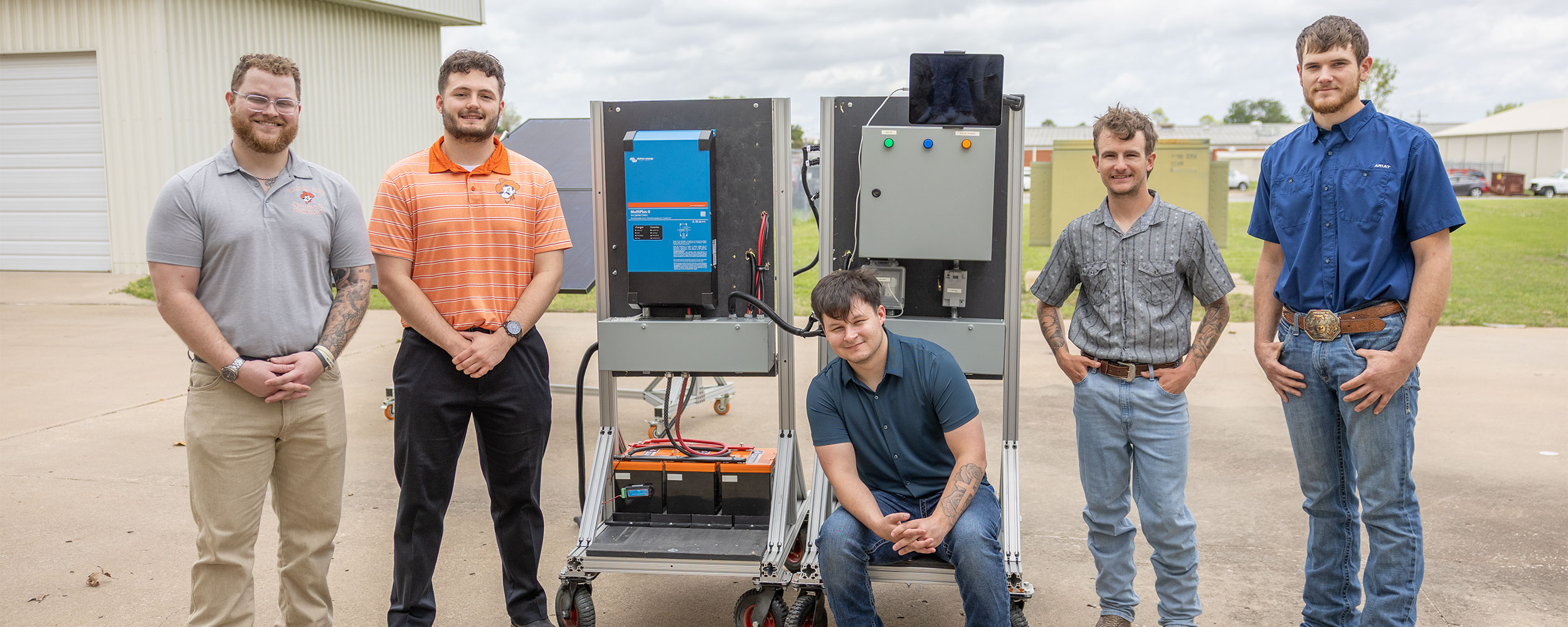 Six people stand outdoors beside two wheeled instrumentation control panels with wiring and solar equipment, posing with a completed technical project in front of campus buildings.