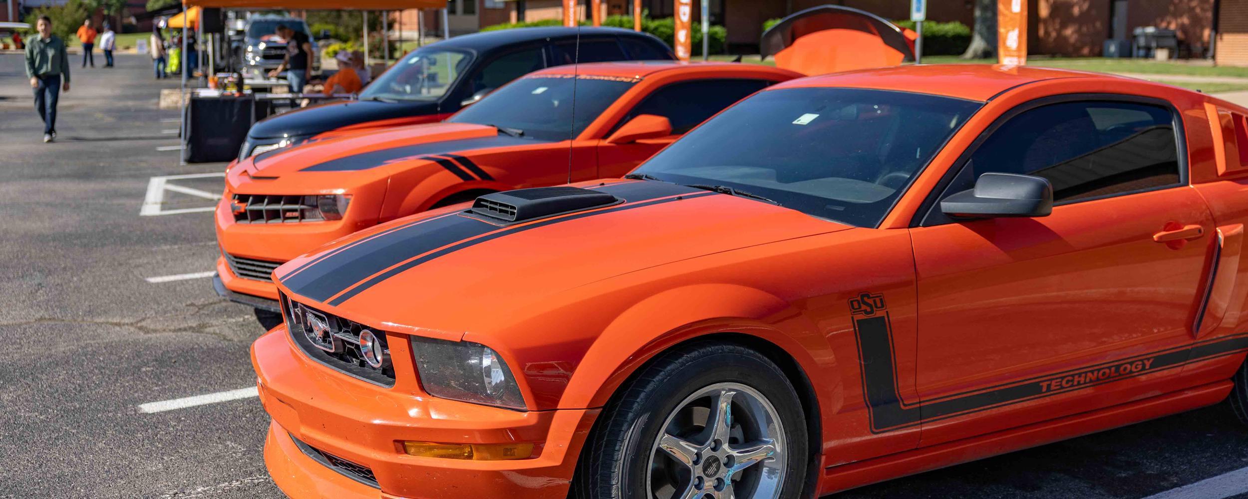 Two bright orange sports cars with black racing stripes are parked side by side outdoors at a campus event, with tents, people, and displays visible in the background.