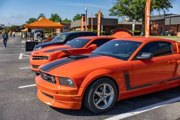 Two bright orange sports cars with black racing stripes are parked side by side outdoors at a campus event, with tents, people, and displays visible in the background.