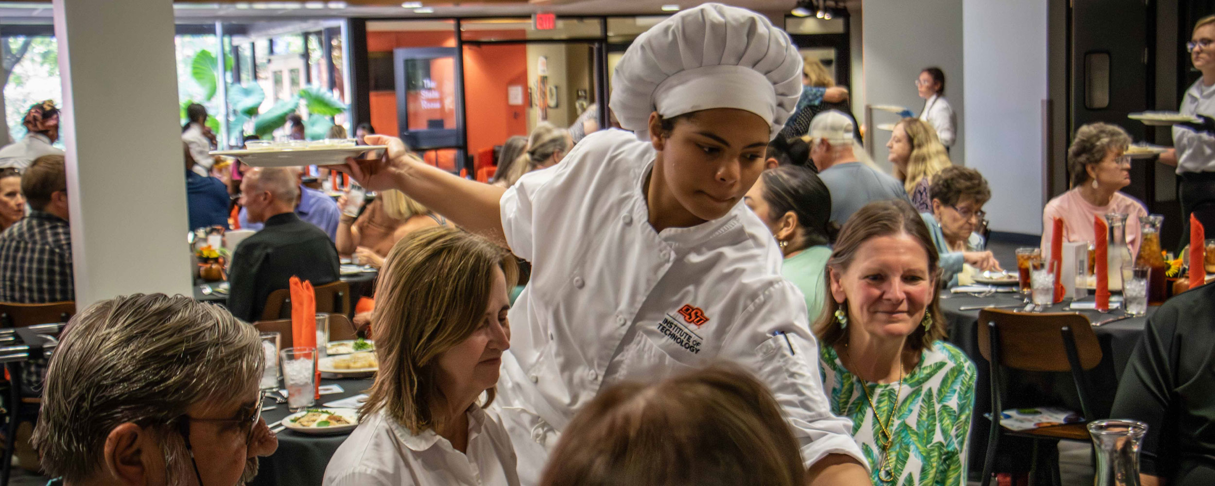 A chef in a white uniform and tall hat serves plated food to seated diners at decorated tables inside a busy restaurant setting, illustrating a student-led culinary service event.