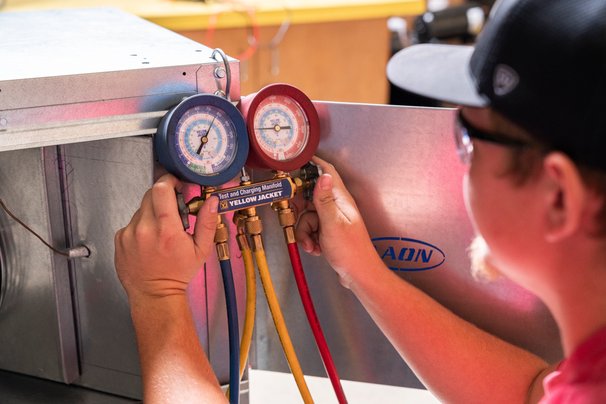 A student in safety glasses checks pressure gauges on an HVAC unit using a Yellow Jacket test and charging manifold.