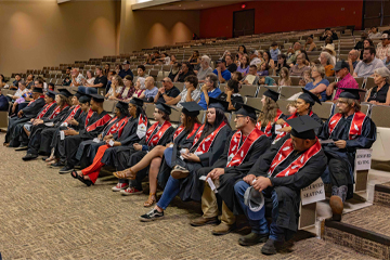 A front row of graduates in caps and gowns sits before a large audience in a tiered auditorium during an OSUIT graduation ceremony. Several graduates are wearing distinctive red stoles with traditional beaded feather designs.