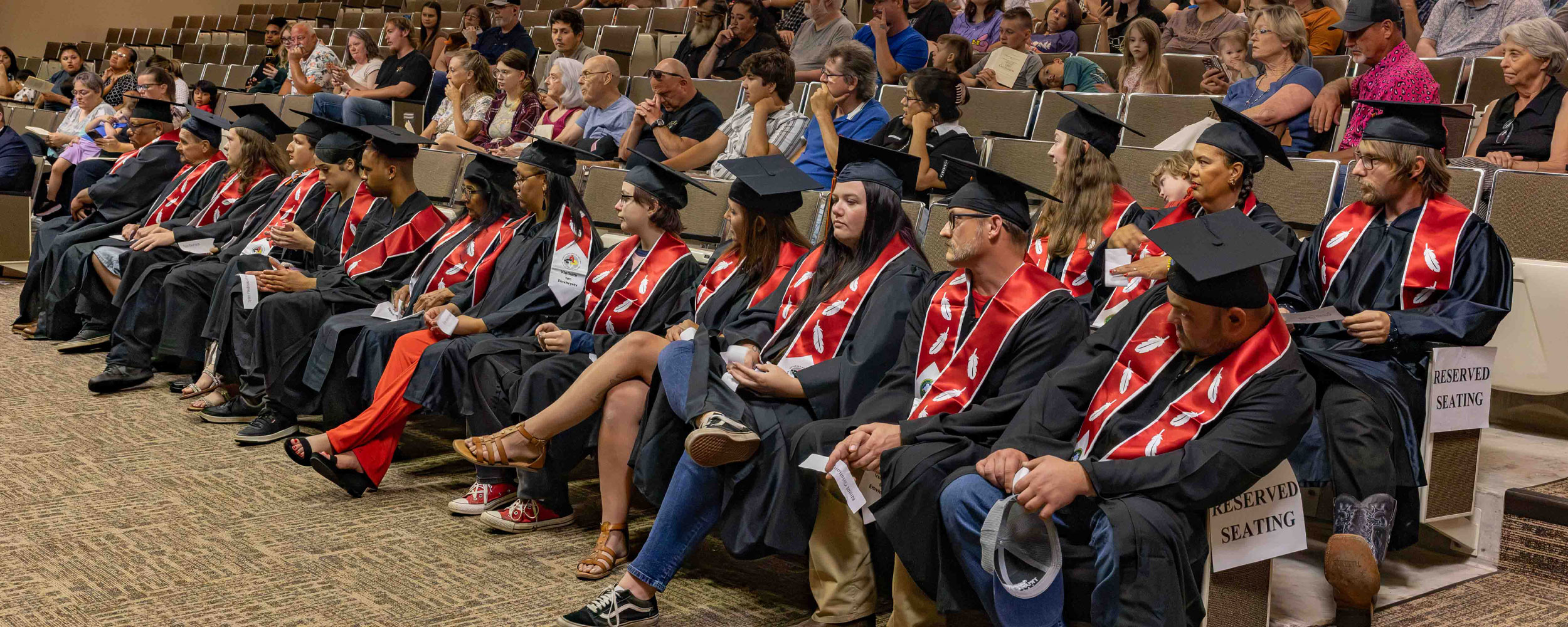 A front row of graduates in caps and gowns sits before a large audience in a tiered auditorium during an OSUIT graduation ceremony. Several graduates are wearing distinctive red stoles with traditional beaded feather designs.