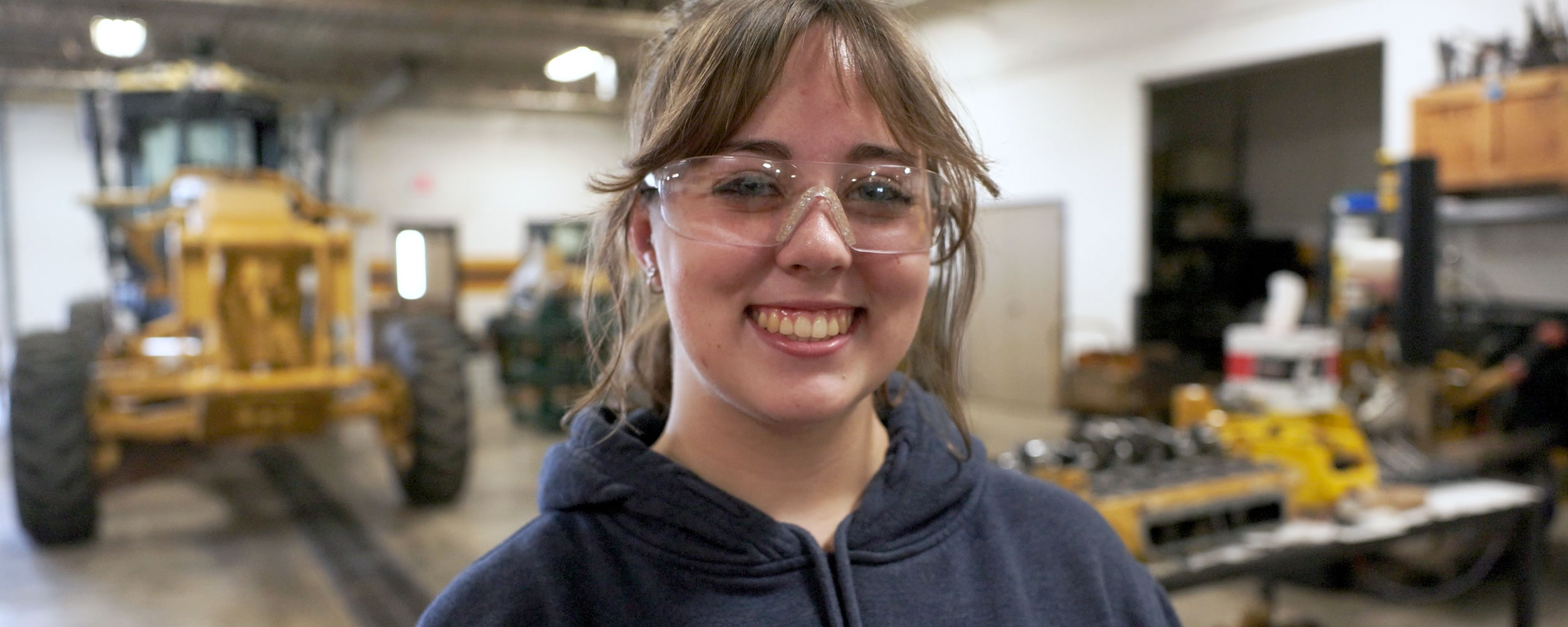 Person wearing clear safety glasses stands in an industrial workshop, with large yellow machinery and tools visible in the background.