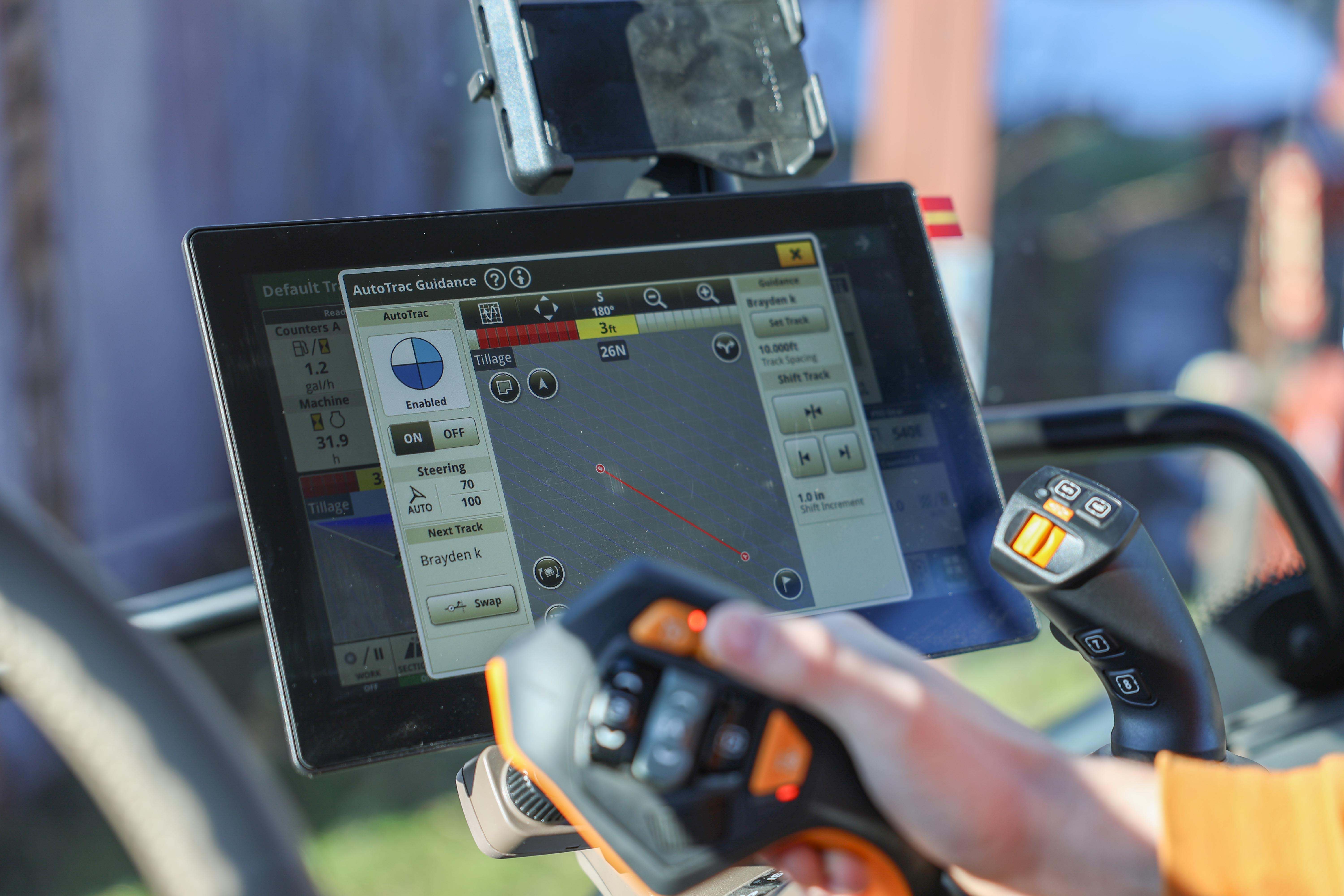 Close-up view of a high-tech tractor control system, featuring a touchscreen display showing AutoTrac guidance settings and a joystick controller with orange and black buttons, used for precision farming operations.