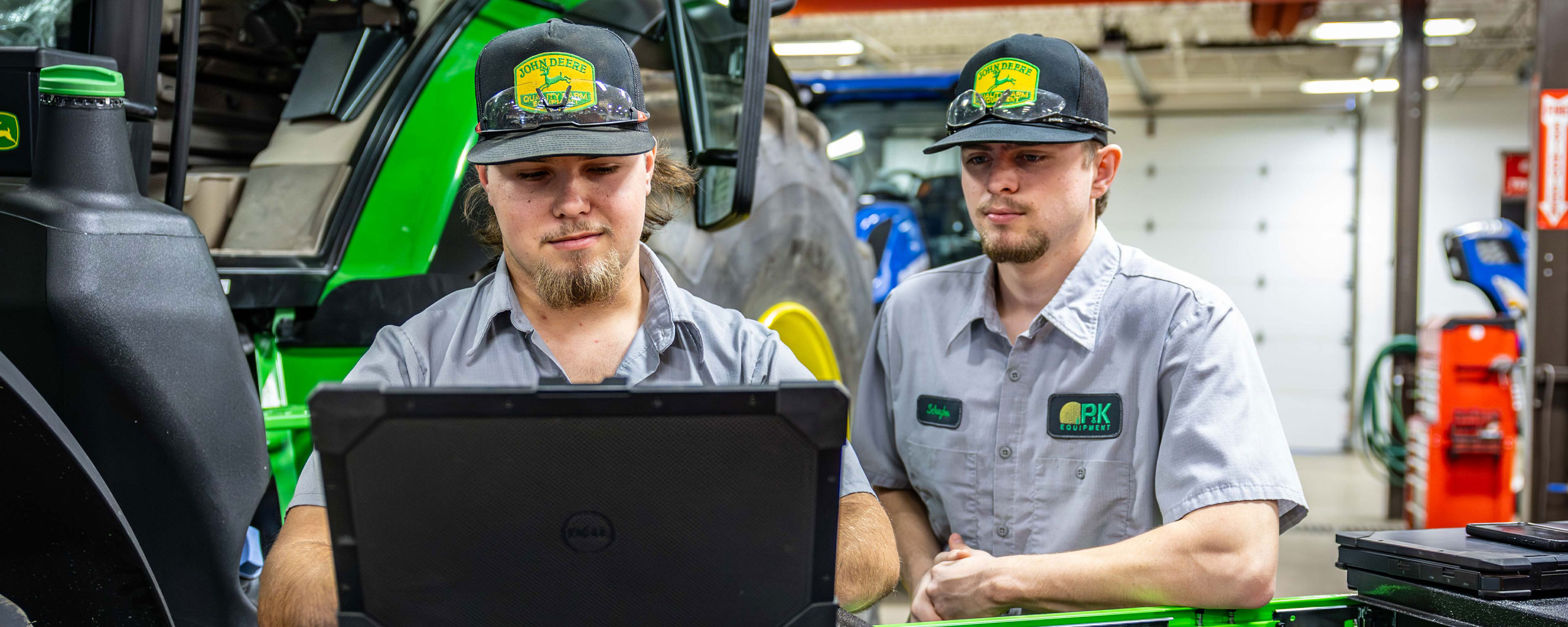 Two technicians wearing matching caps and work shirts stand in a workshop beside a large green tractor, focusing together on a laptop as part of hands-on equipment diagnostics or training.