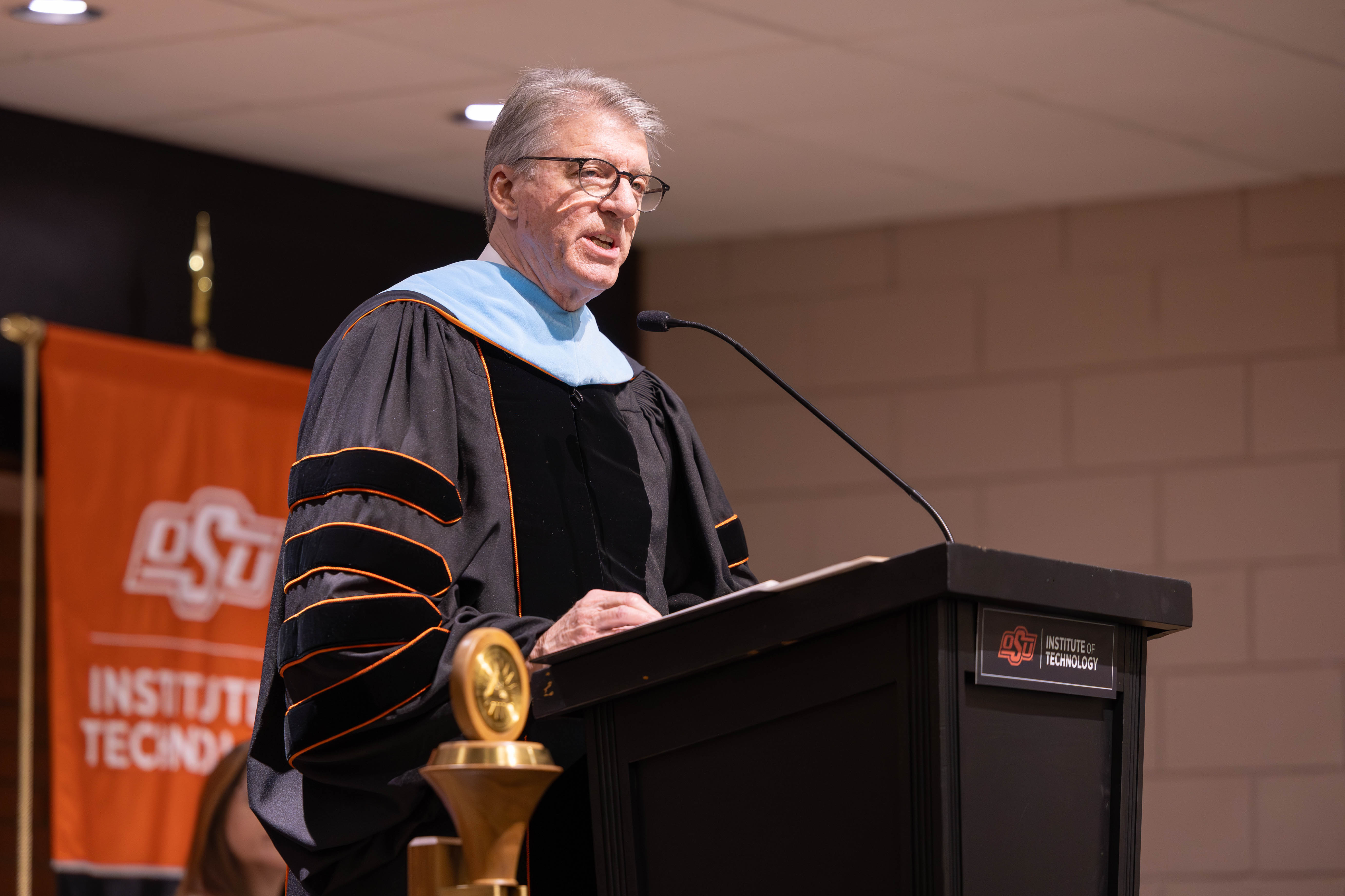 A speaker in academic regalia stands at a podium delivering remarks during an OSU Institute of Technology commencement ceremony, with an OSU banner and ceremonial mace visible nearby.