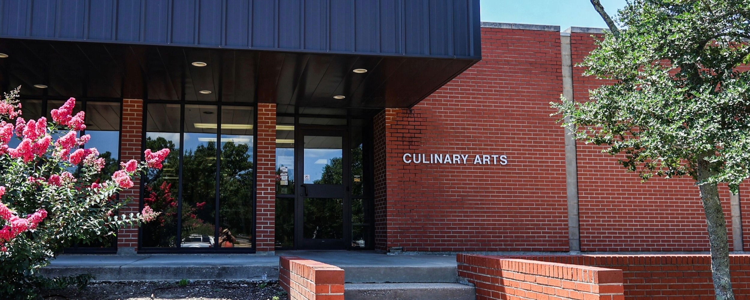 The exterior of a red brick building labeled “Culinary Arts” features a glass entrance, landscaped plants, and a shaded overhang, identifying the facility as a culinary arts academic building.