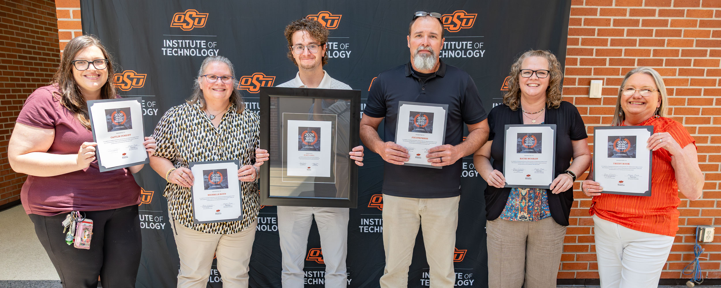 Six smiling OSUIT faculty and staff members stand in a line holding their 2026 Faculty Staff Awards certificates and one large framed award. They are posing in front of a black Oklahoma State University Institute of Technology backdrop and a brick wall.