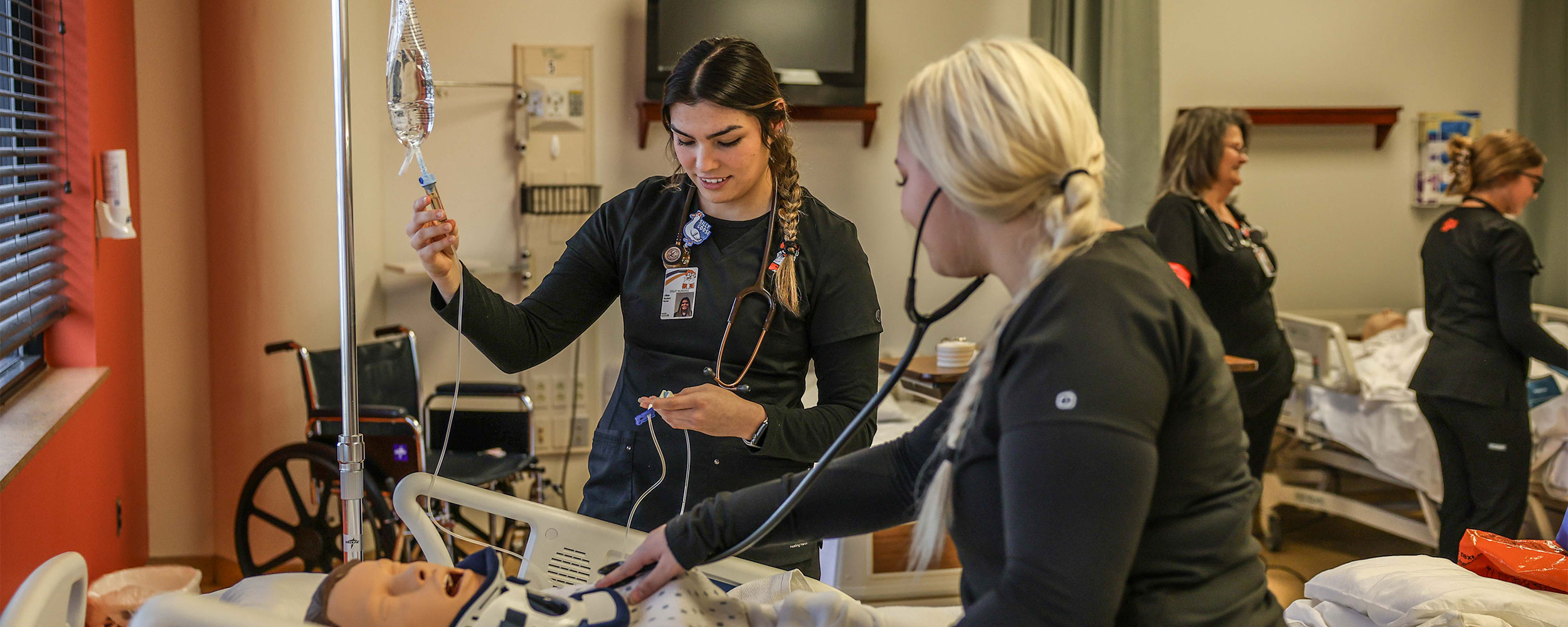 OSUIT nursing students in black scrubs practice clinical skills on a medical mannequin in a simulation lab.