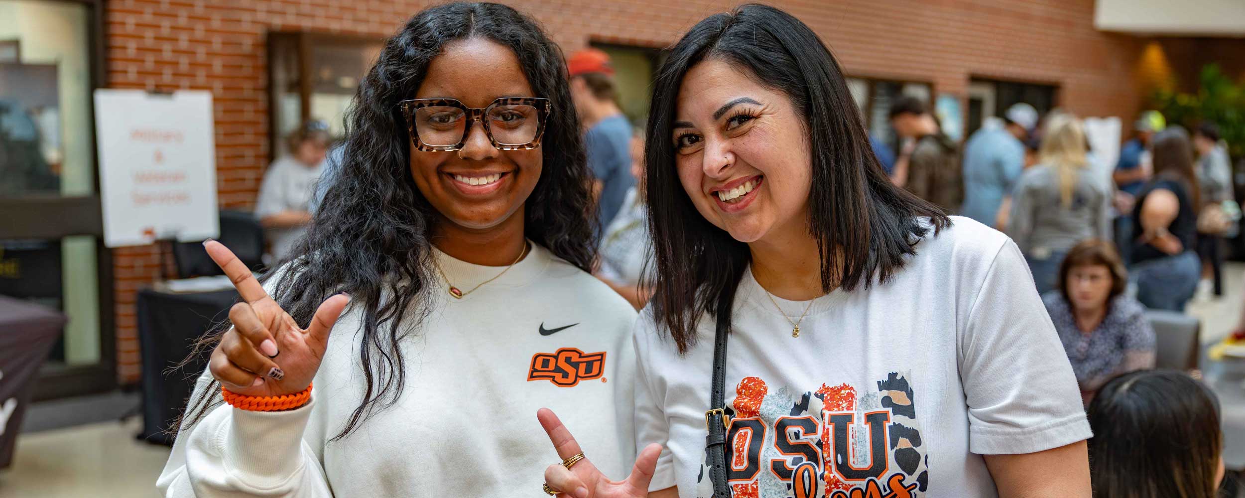 Two women at a campus event, one with dark wavy hair and clear glasses flashing the OSU sign, and the other with shoulder-length dark hair and a wide smile, wearing OSU apparel. They are indoors with a brick wall and a diverse crowd in the background.