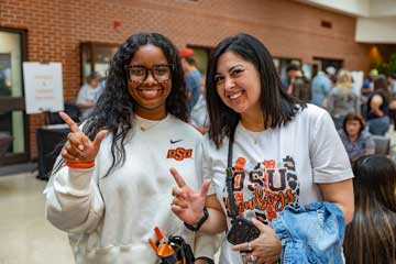 Two women at a campus event, one with dark wavy hair and clear glasses flashing the OSU sign, and the other with shoulder-length dark hair and a wide smile, wearing OSU apparel. They are indoors with a brick wall and a diverse crowd in the background