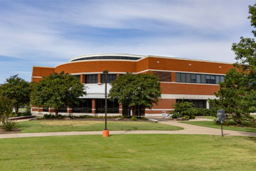 Exterior view of the Donald W. Reynolds Center at OSUIT, a two-story brick building with curved architectural facade, surrounded by trees and walking paths.