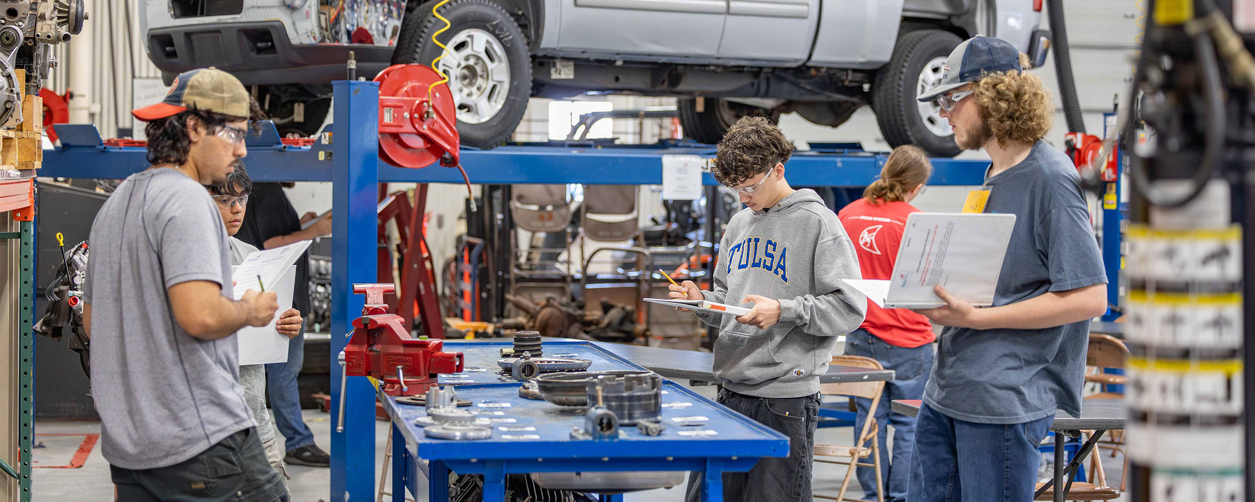Students wearing safety glasses work at a bench in an automotive training lab, reviewing checklists and components while vehicles are raised on lifts in the background, illustrating hands-on technical education.
