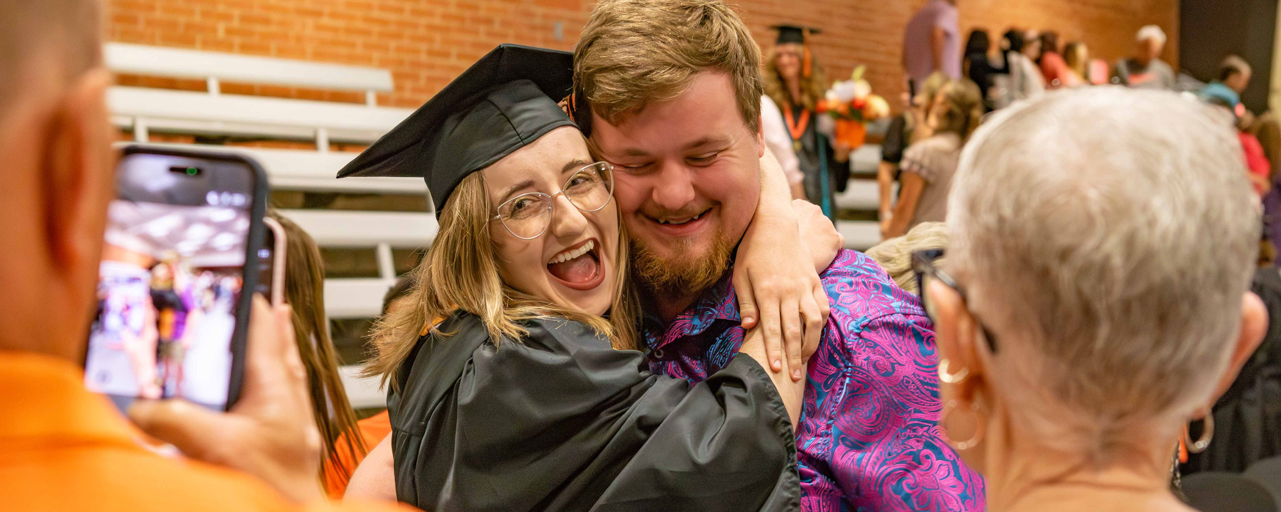 A graduate wearing a cap and gown hugs another person during a commencement ceremony while others nearby watch and take photos inside a gymnasium or event hall.