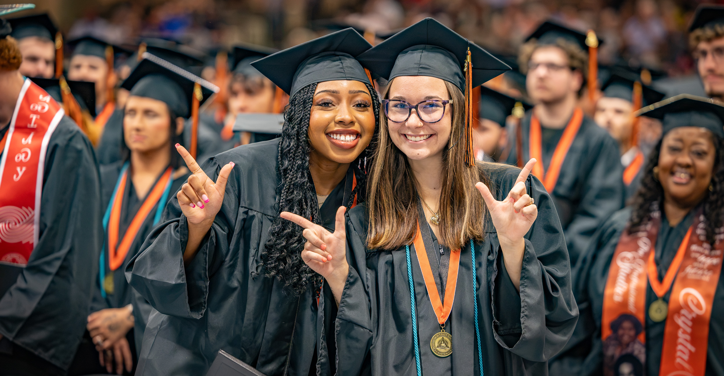 Two OSUIT graduates in black caps and gowns smile at the camera and pose with hand gestures while seated among fellow graduates during a commencement ceremony.
