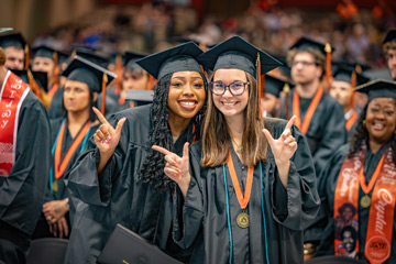 Two OSUIT graduates in black caps and gowns smile at the camera and pose with hand gestures while seated among fellow graduates during a commencement ceremony.