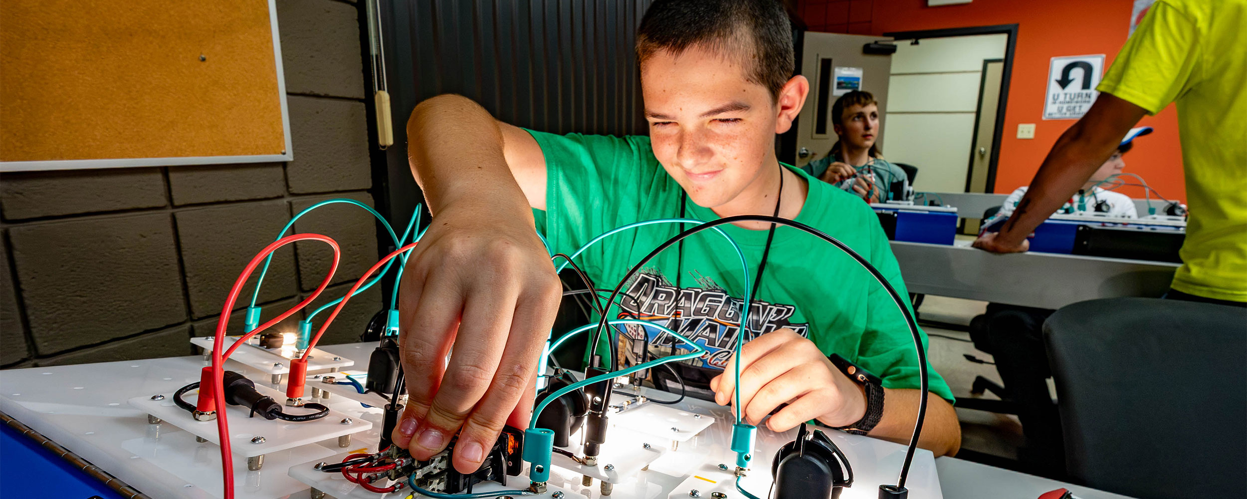 Student works hands-on with an electrical circuit training board, connecting wires and components during a STEM activity at OSUIT Summer Academy while other participants use similar equipment in a classroom setting.