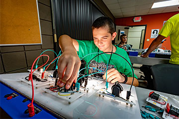 Student works hands-on with an electrical circuit training board, connecting wires and components during a STEM activity at OSUIT Summer Academy while other participants use similar equipment in a classroom setting.