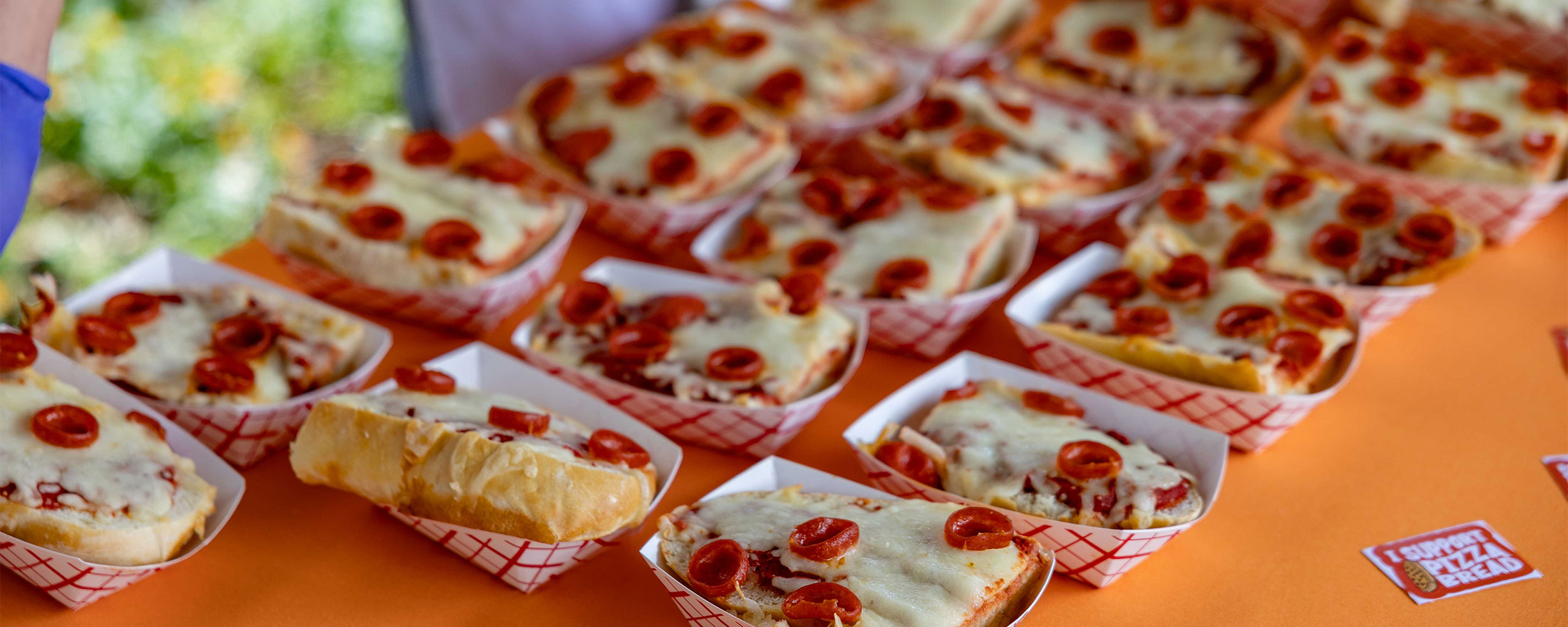 Rows of rectangular slices of pizza bread topped with melted cheese and pepperoni are displayed in paper trays on an orange table at a food event or gathering.
