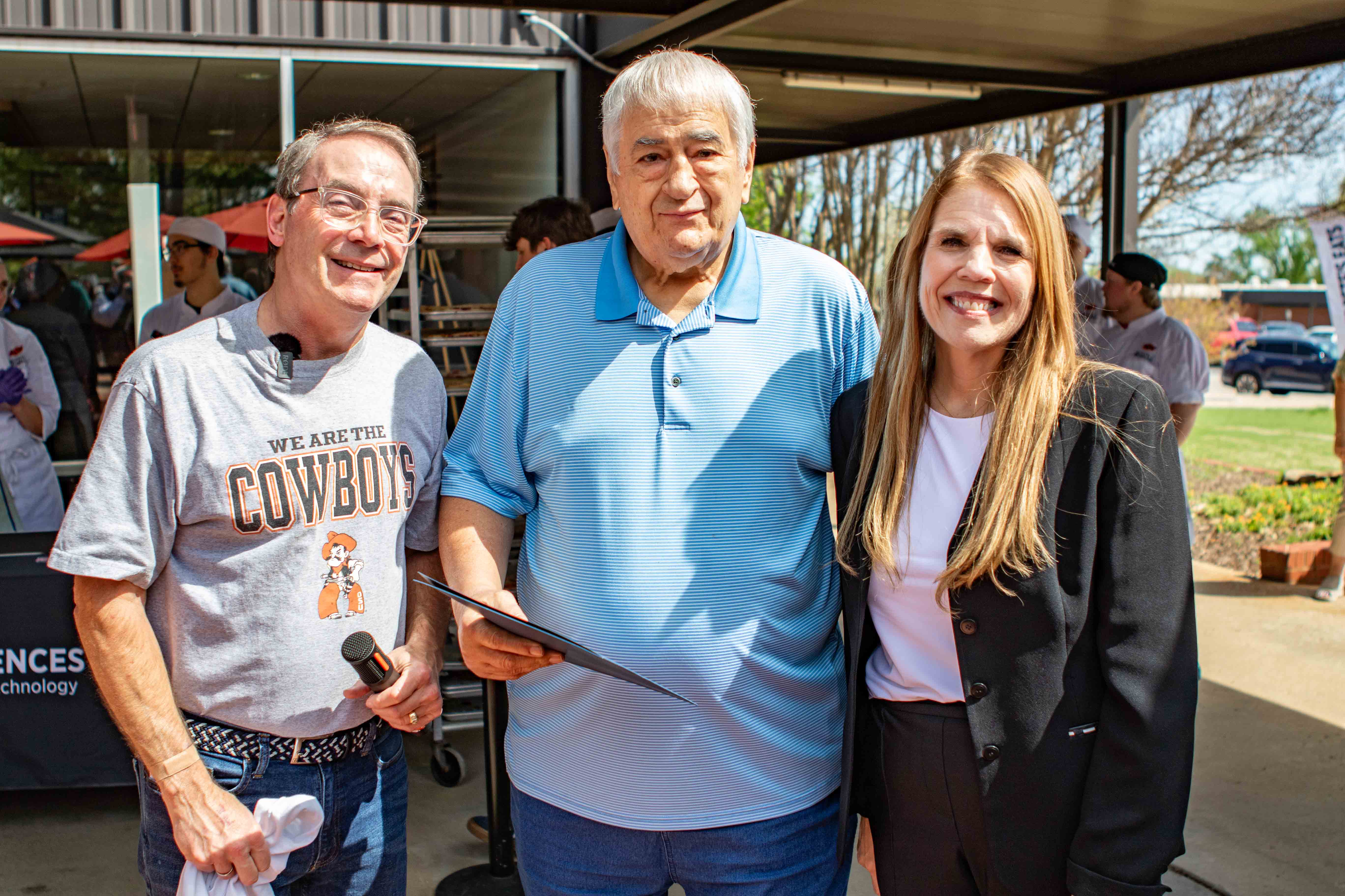 Three adults stand together outdoors at a campus event, with one holding a microphone and another holding a document, posing in front of a building and event signage.