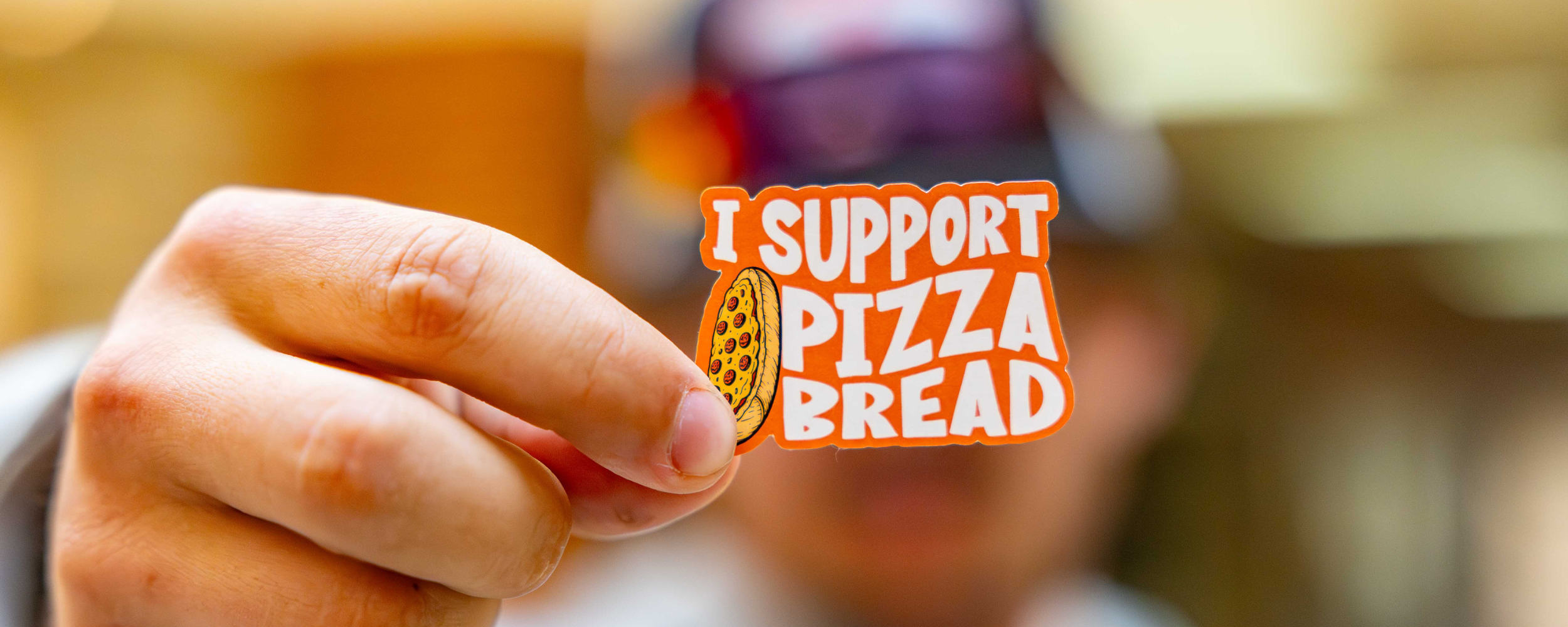 A hand holds a small sticker reading ‘I support pizza bread,’ featuring a cartoon slice of pizza bread against a softly blurred background, suggesting a playful food‑themed message.
