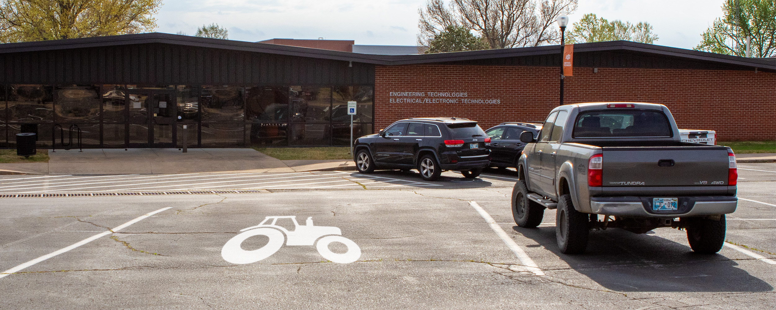 A brick engineering technologies building is shown with a parking lot in front featuring a marked tractor parking space and several parked vehicles, illustrating facilities designed to accommodate agricultural or technical programs.