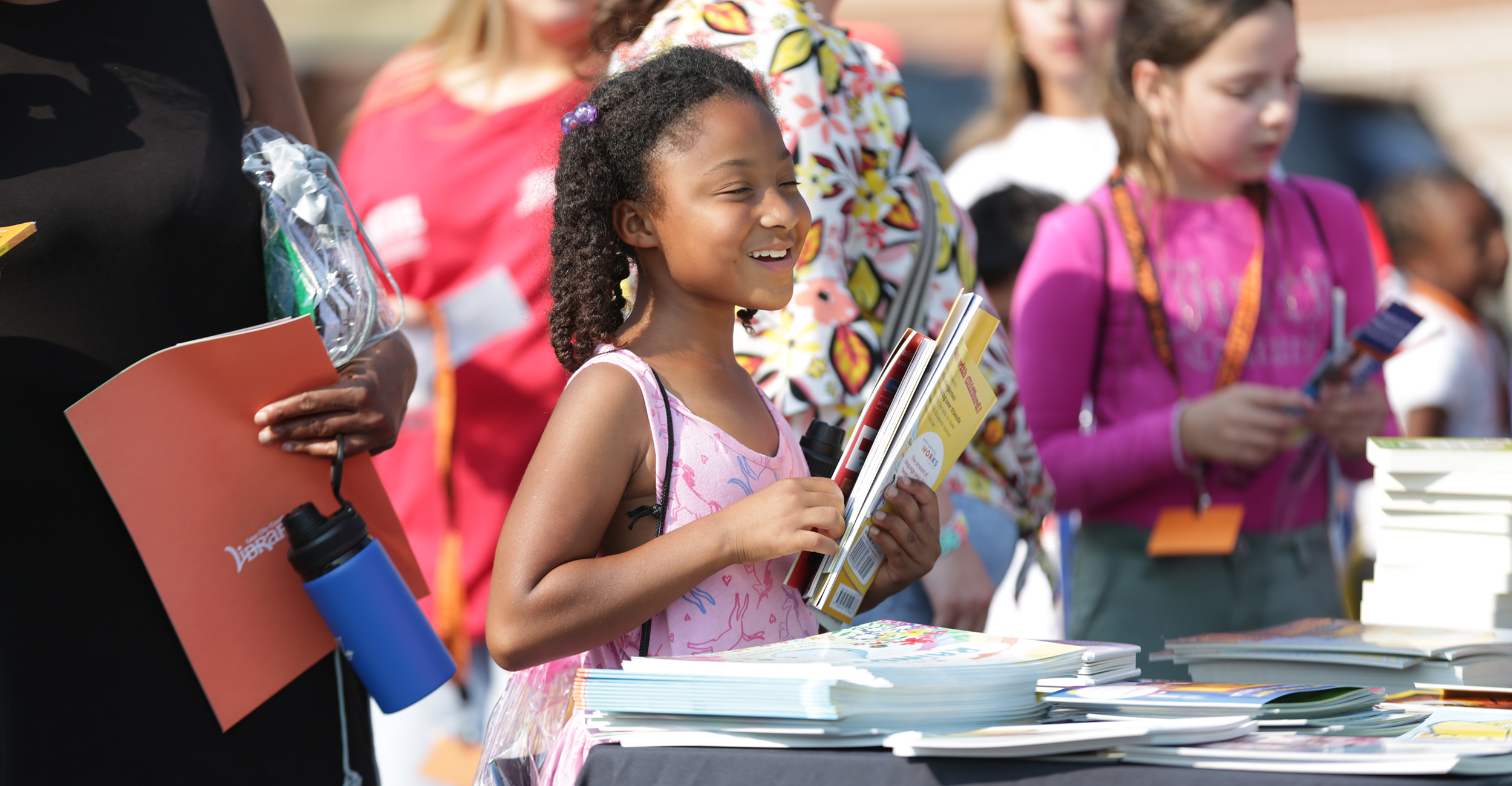 A girl standing in front of a table full of books, smiles and holds up a stack of notebooks, surrounded by a crowd of eventgoers at the 2026 OSU-Tulsa Back to School Bash.