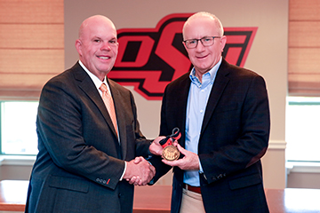 Two individuals in business attire shake hands while one presents a medallion in front of an Oklahoma State University logo inside a formal indoor setting.