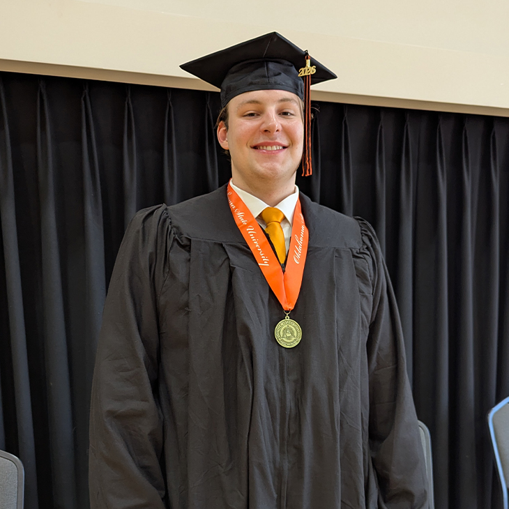 A graduate wearing a black cap and gown with an orange stole and medal stands in front of a dark curtain backdrop, posing for a formal graduation portrait.