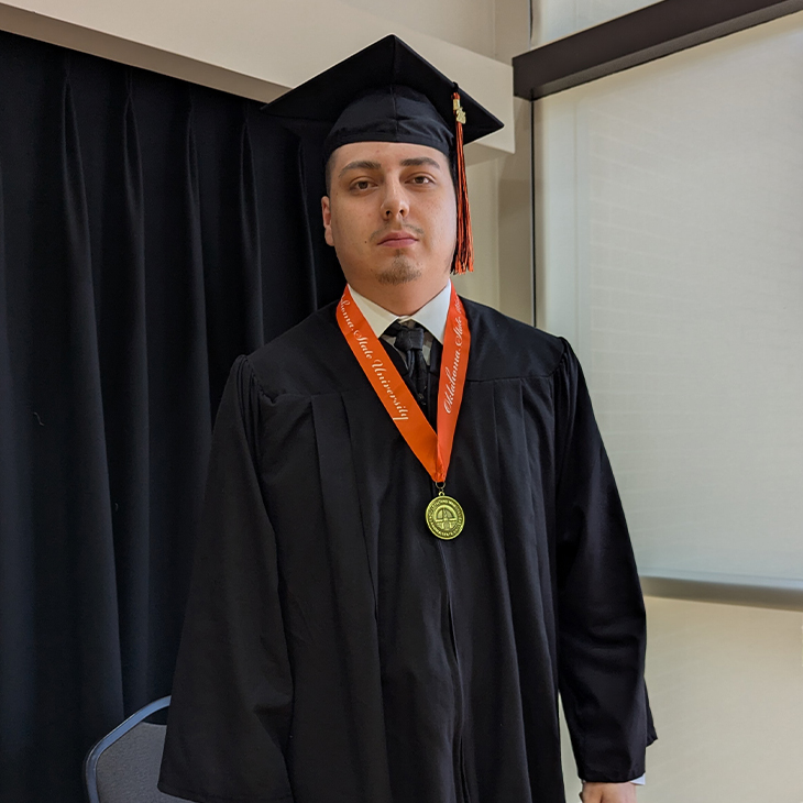 A graduate wearing a black cap and gown with an orange honor stole and medal stands indoors near a window and dark curtain, posing for a formal graduation portrait.
