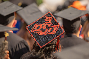 A crowd of graduates wearing black caps and gowns is seen from behind, with one decorated mortarboard featuring the OSU logo and the number “26” prominently centered, indicating a graduation ceremony.