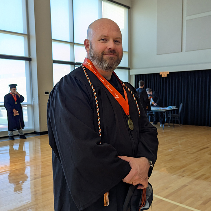 A person wearing a black graduation gown with an orange honor stole and medal stands inside a gymnasium, posing for a portrait while others in academic attire gather in the background near tables.