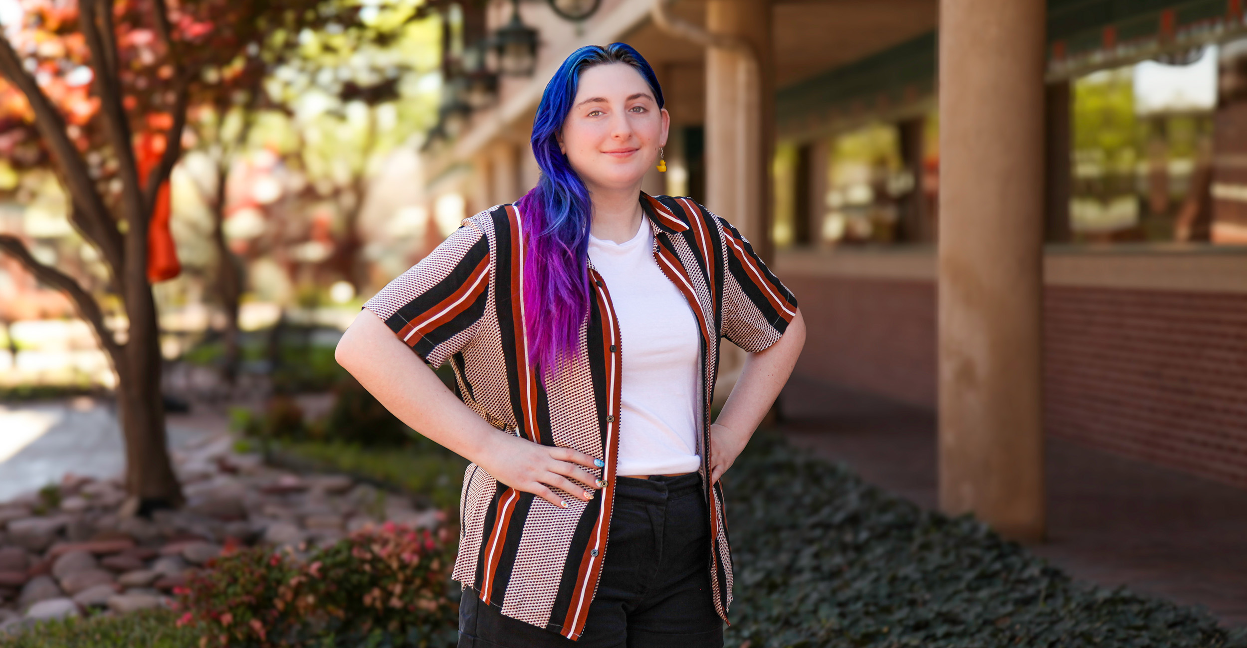 Person with long blue‑and‑purple hair stands outdoors on a campus walkway with hands on hips, posed for a portrait with trees and a university building in the background.