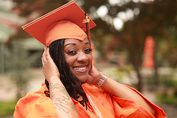 An individual wearing an orange graduation cap and gown poses outdoors on a campus setting, holding both hands near the sides of the mortarboard for a close-up graduation portrait.
