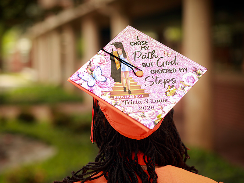 The back view of a graduation cap decorated with floral designs and the message “I chose my path, but God ordered my steps,” along with a scripture reference and graduation year, worn during an outdoor graduation photoshoot.