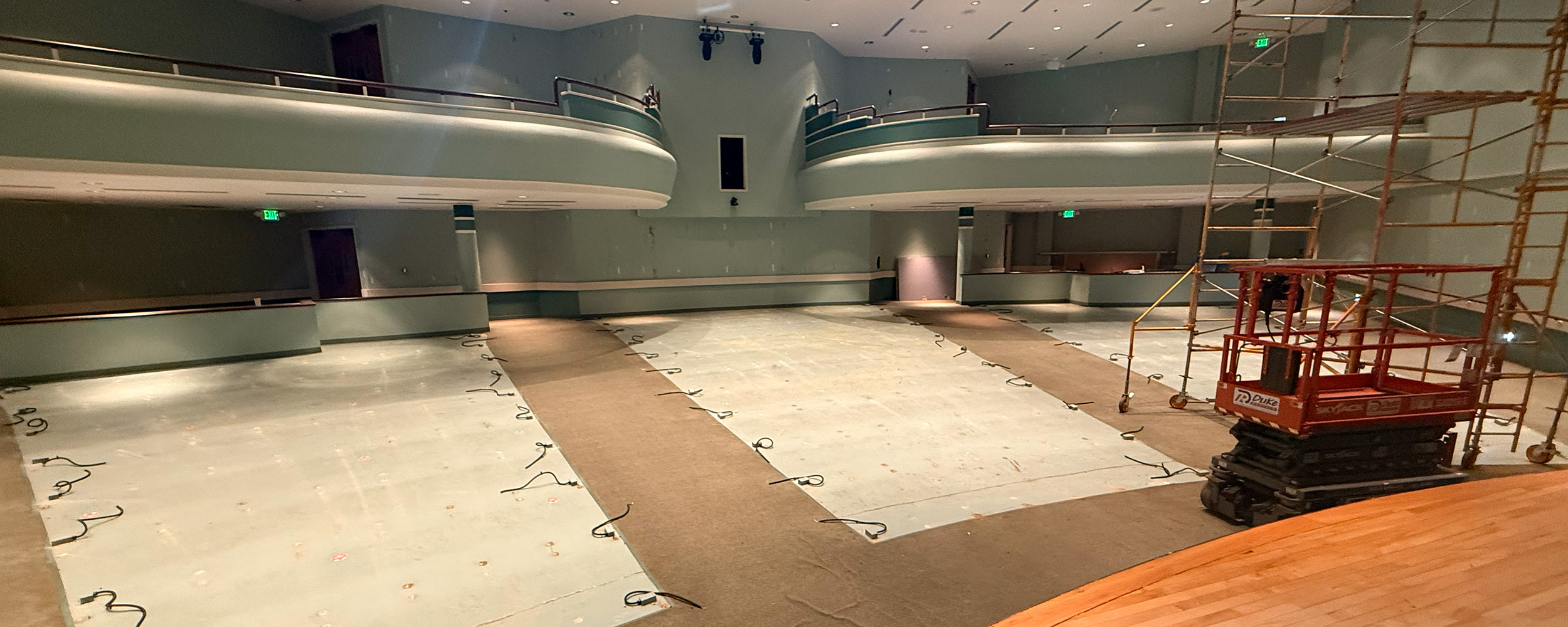 The OSU-Tulsa Auditorium space, now emptied of chairs, is visibly under construction. The photo, taken from the stages, shows rows of white flooring where seats used to be, surrounded by carpet, with scaffolding on the righthand side of the image.