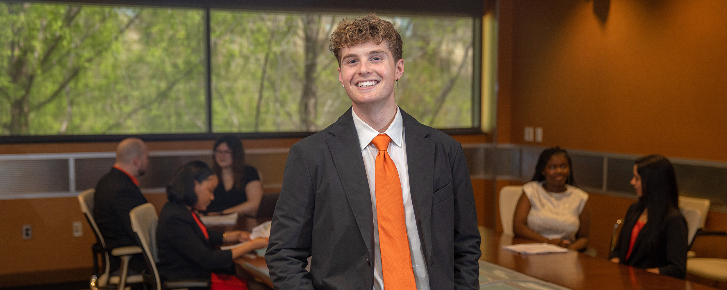 A portrait of a College of Professional Studies student in a suit and orange tie, posed in a conference room with professionals in the background.