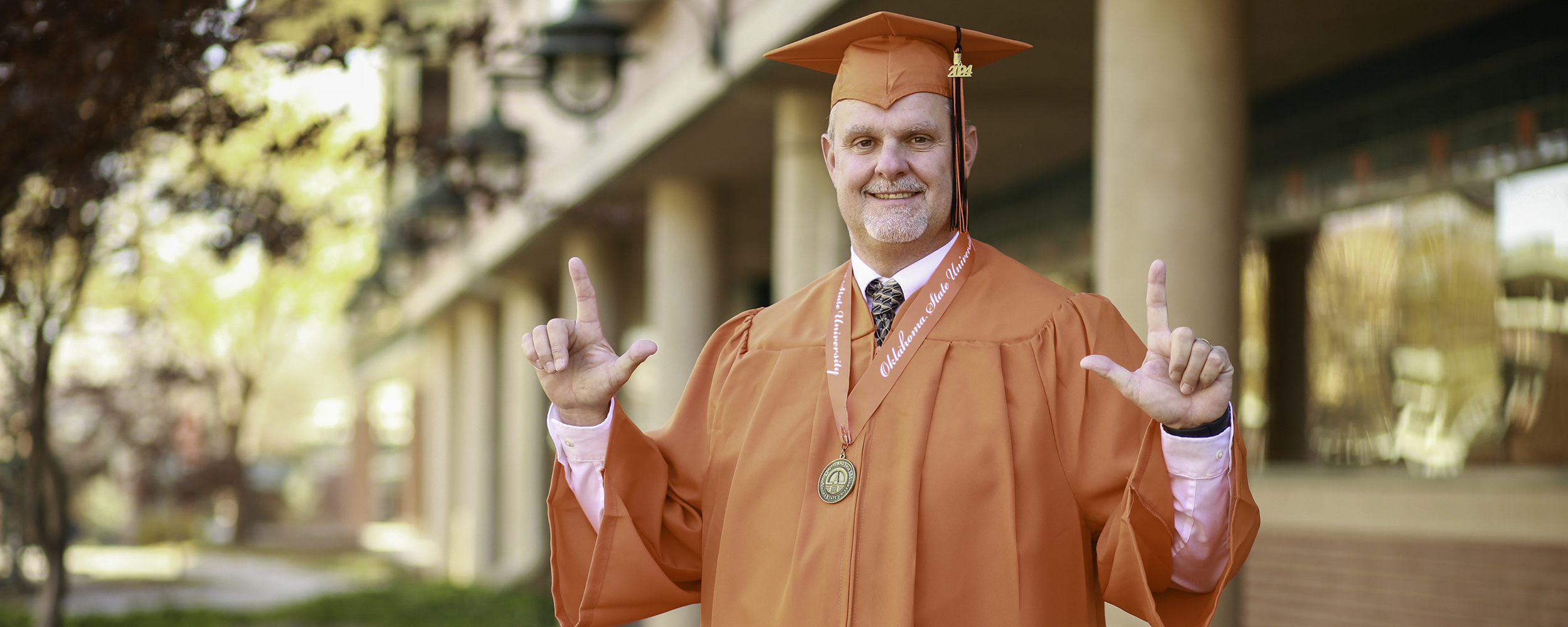 Fall 2024 College of Professional Studies Orange Gown Graduate Dallas Smith poses in the OSU-Tulsa courtyard in his orange gown, sporting "Go Pokes" finger pistols.
