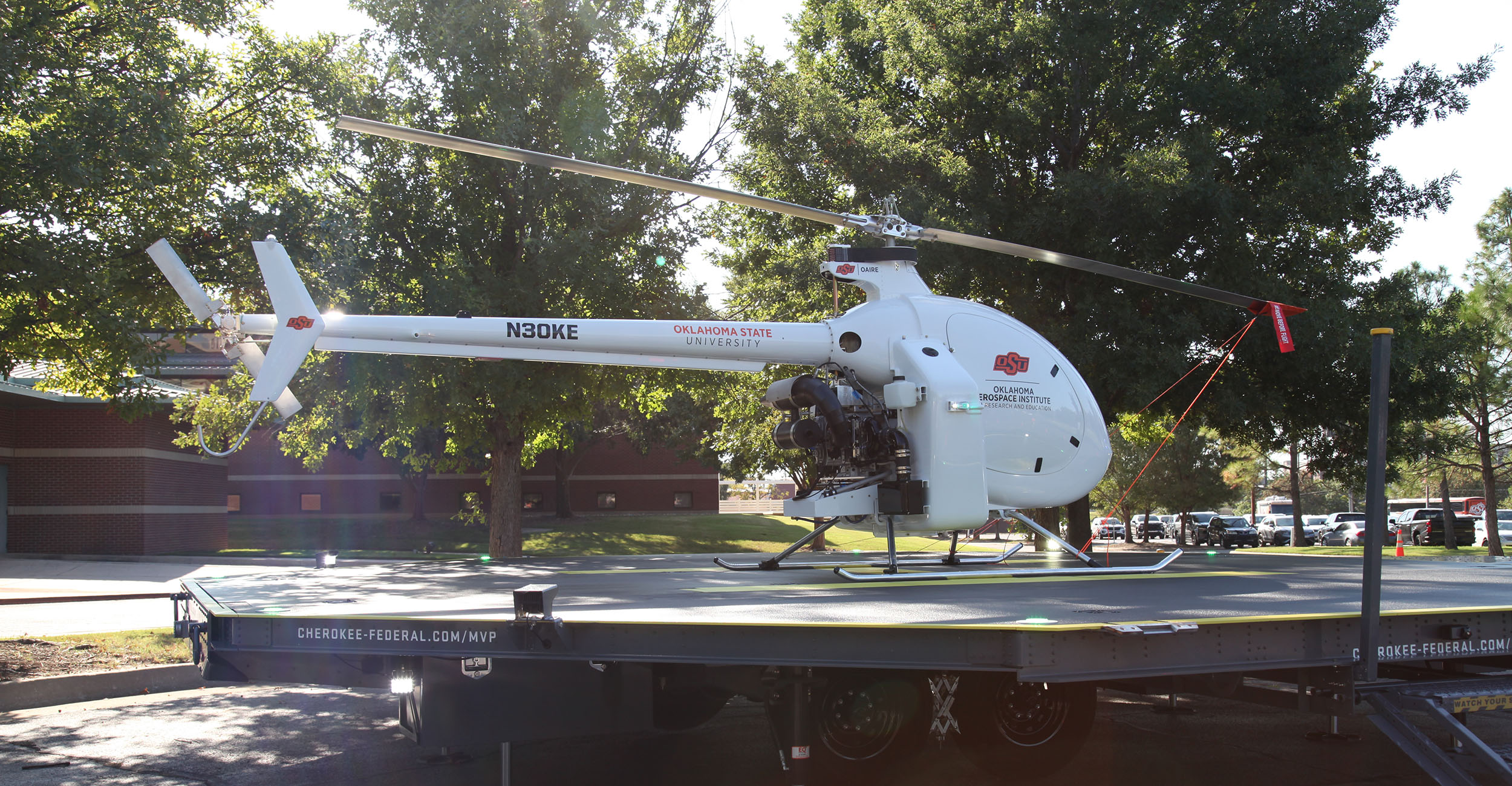 A large autonomous aircraft on display outside the Main Hall Building on the OSU-Tulsa campus. it is mounted on a small helipad, is sleek and white with propellors like a helicopter, and features Pistol Pete's face on the front where a cockpit would be.