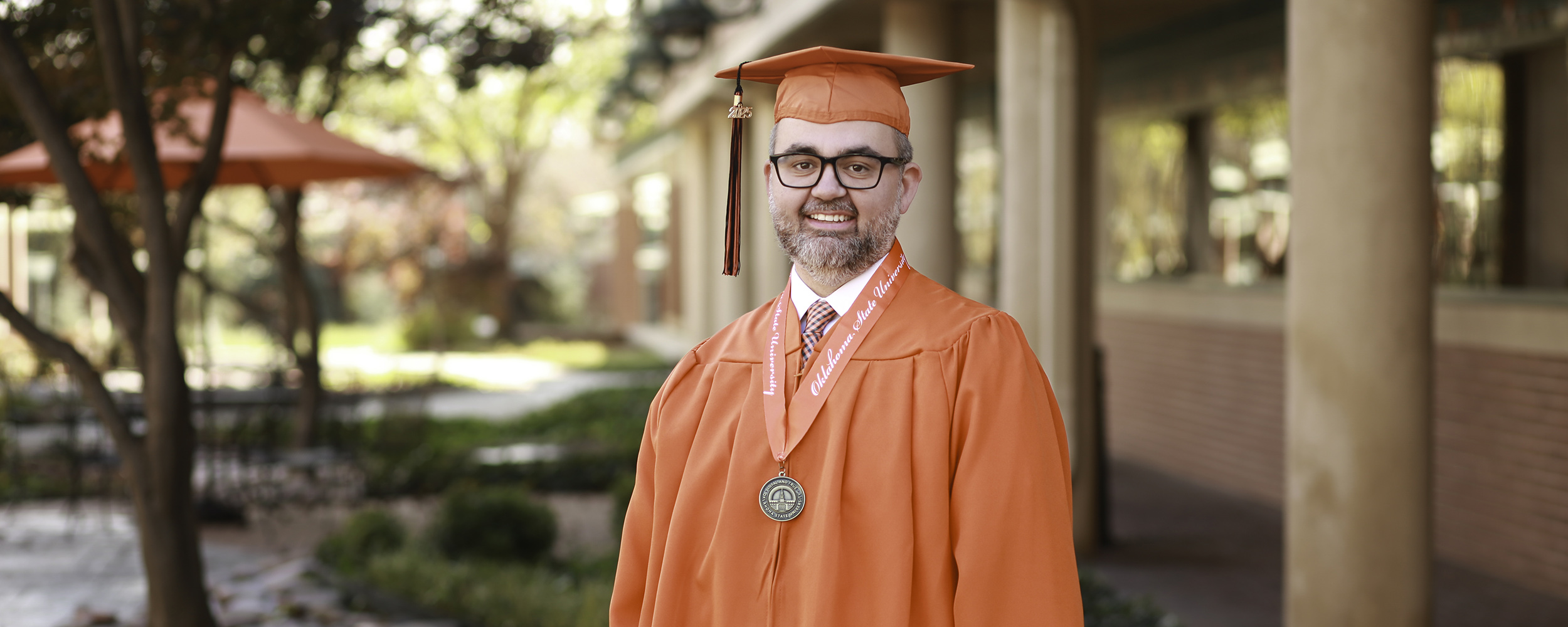 Stephen Taylor poses in his orange cap and gown in a shallow depth of field shot in the OSU-Tulsa Courtyard, focused on Taylor in the foreground and offering a blurry background of tan columns, red brick and leafy fall foliage.