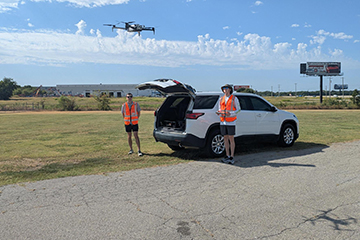 Two pilots in orange safety vests fly a drone in a Tulsa parking lot.