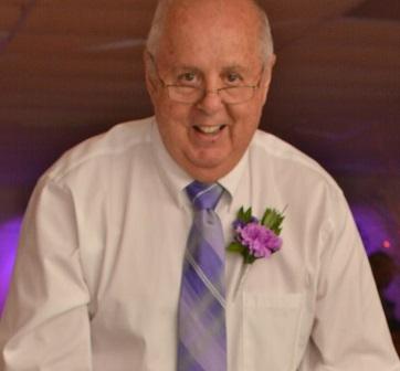 An older man sits with a white shirt, purple flower and purple tie.