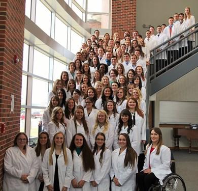 Dozens of students wearing white coats pose on a staircase.