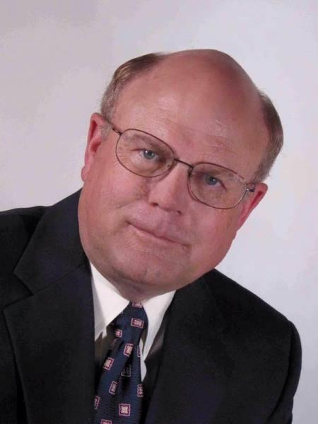A balding Caucasian man wearing a black suit and tie poses for a picture.