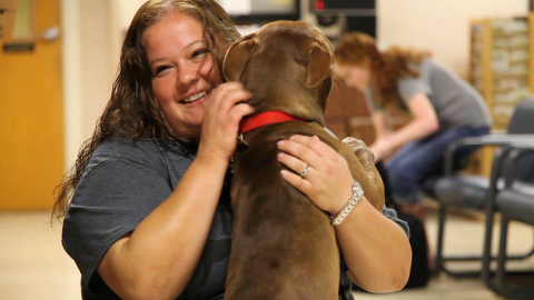 A woman smiles as she hugs a brown dog.